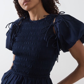 Model wearing Damson Madder navy cotton blouse with puff sleeves, shirred bodice and tie bow details at shoulders.