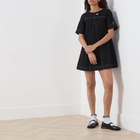 Model wearing Damson Madder black mini dress with contrast white stitching and short sleeves, styled with white socks and black Mary Jane shoes.
