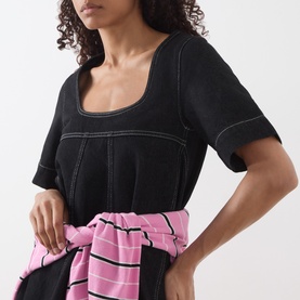 Model wearing Damson Madder black cotton dress with contrast stitching, styled with a pink striped shirt tied at the waist.