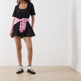 Model wearing Damson Madder black mini dress with pink tied scarf at waist, styled with white socks and black loafers.