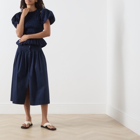 Model wearing Damson Madder navy cotton midi skirt with elasticated waist and front split, styled with matching navy top and tan sandals.