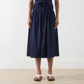 Model wearing Damson Madder navy cotton midi skirt with smocked waistband and tie detail, styled with white strappy sandals.