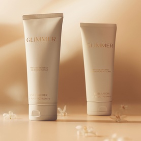 Est&eacute;e Lauder Glimmer body lotion and cream duo in beige tubes, styled on a warm neutral background with delicate floral accents.