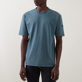 Model wearing Belstaff Crew Neck T in petrol blue cotton, with small arm badge detail, styled with dark jeans.