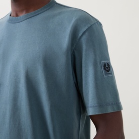 Model wearing Belstaff Crew Neck T-Shirt in petrol blue cotton, showing sleeve badge logo detail at close range.