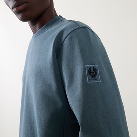 Model wearing Belstaff petrol cotton sweatshirt, close-up of sleeve showing woven logo badge detail.