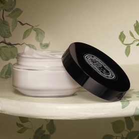 Diptyque body cream in open frosted glass jar with black lid, white cream texture visible, styled with botanical backdrop