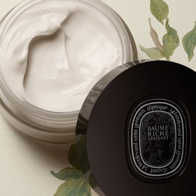Diptyque Baume Riche Apaisant body cream in open black jar, revealing thick white cream texture, with botanical leaves in background