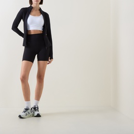Model wearing On Running black zip-up jacket with white sports bra, black cycling shorts, and chunky white trainers.