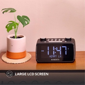 Roberts Ortus 4 FM Dual Alarm Clock Radio in black, showing large blue LCD screen displaying time, on wooden surface beside potted plant