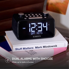 Roberts Ortus 4 FM Dual Alarm Clock Radio in black, displaying time 12:34 on white LED screen, shown on stacked books on bedside table.