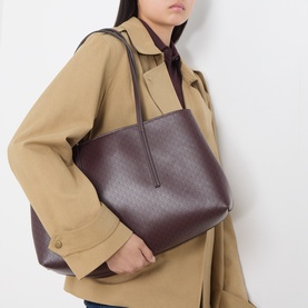 Model carrying Calvin Klein Emblem Medium Work Tote Bag in Fudge AOP with all-over print and twin shoulder straps, styled with a beige trench coat.