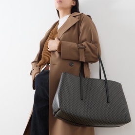Model carrying Calvin Klein Emblem Medium Work Tote Bag in black monogram print with dual shoulder straps, styled with a tan trench coat