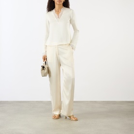 Model wearing Ganni egret long-sleeve V-neck blouse with wide-leg trousers, carrying a grey handbag, styled with gold sandals