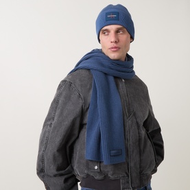 Model with Calvin Klein Patch Chunky Ribbed Logo Scarf in Dark Denim, ribbed knit with logo tag, styled with matching beanie and grey jacket