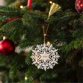 Villeroy & Boch white lattice snowflake Christmas tree ornament with gold tassel, hanging on a decorated green tree with red baubles.