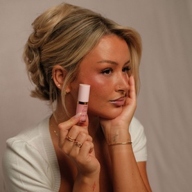 Model with Kash Beauty Skin Glaze blush, holding pink mini bottle, wearing white top, rings and gold bracelet