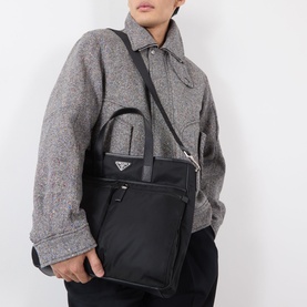 Model carrying Prada black recycled polyamide tote bag with silver triangle logo, top handles and crossbody strap.