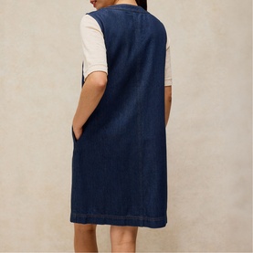 Model wearing White Stuff dark denim pinafore dress, styled over a white short-sleeve top, shown from behind, knee-length with side pockets.