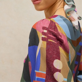 Model wearing White Stuff dress in pink multicolour abstract print, styled with a printed hair scarf and delicate necklace.