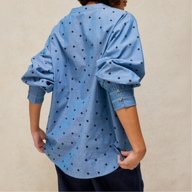 Model wearing White Stuff blue multi spot print oversized shirt, back view, with gold button cuffs, styled with navy trousers.