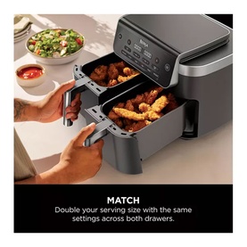 Ninja dual-drawer air fryer in grey, open with cooked food visible, hands pulling out drawer, salad and dipping sauce in background