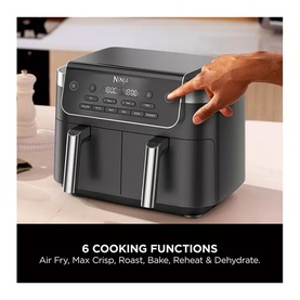 Ninja dual-basket air fryer in dark grey with digital display panel, hand shown pressing controls, text listing 6 cooking functions below