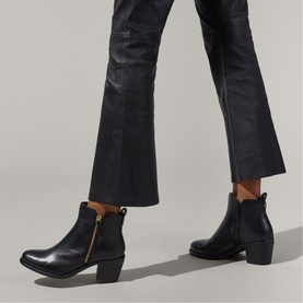 Model wearing Carvela black leather ankle boots with gold zip detail and block heel, styled with cropped flared black trousers.