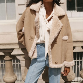 Model wearing Mint Velvet brown shearling-trim jacket styled with white blouse and light blue jeans, shot outdoors.