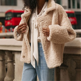Model wearing Mint Velvet brown teddy fleece jacket, styled with white tie-neck blouse and light blue jeans.