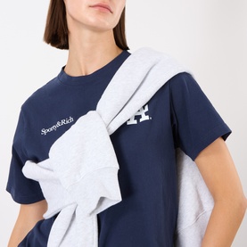 Model wearing Sporty and Rich Dark Navy T-Shirt with logo print, styled with a light grey sweatshirt tied over the shoulder.