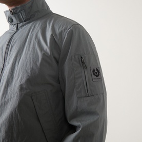 Model wearing Belstaff Casual Jacket in Atlantic Grey, close-up of sleeve badge logo and zip chest pocket detail.