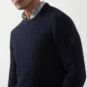 Model wearing Belstaff Dark Ink cable-knit crew neck jumper, styled over a checked shirt, with logo badge detail on sleeve.