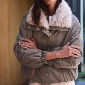 Model wearing Mint Velvet green padded jacket with faux fur collar, gold snap buttons and elasticated cuffs, arms crossed.