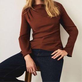 Model wearing rust brown long-sleeve top with asymmetric neckline and flared cuffs, styled with dark navy jeans