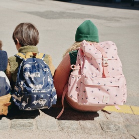 Two children seated outdoors, one carrying Liewood backpack in cherry heart rosey pink print, second child wearing navy printed backpack