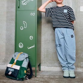 Child leaning against green locker wearing striped tee and grey joggers, with Liewood play multi mix backpack on floor beside them