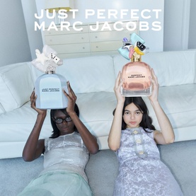 Two models holding oversized Marc Jacobs Just Perfect Eau de Parfum bottles in blue and pink, lying on a white sofa
