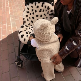 BiniBamba Leopard pram liner in use on a pushchair, with toddler in cream teddy bear suit standing alongside