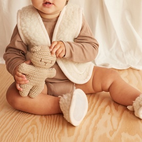 Baby wearing BiniBamba Milk soft sole booties, seated holding a crochet teddy, styled with a taupe romper and cream gilet
