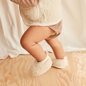 Model wearing BiniBamba Milk shearling baby booties, styled with matching cream teddy coat and shorts, on wooden floor