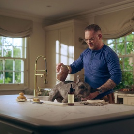 Man washing a grey French Bulldog on a kitchen counter beside a Jo Malone shampoo bottle, with a scrubbing brush nearby.