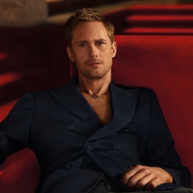 Model with Valentino Vendetta Uomo Eau de Parfum, seated in navy double-breasted suit, gold chain necklace, red ambient setting