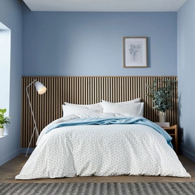 White ditsy floral duvet set styled on a double bed with light blue throw, wooden headboard panel, and floor lamp in a blue-toned bedroom