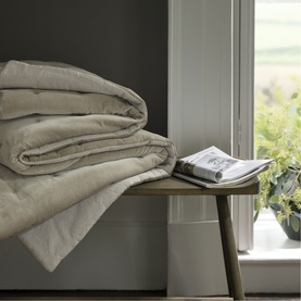 Neatly folded natural linen duvet stacked on a wooden bench beside an open magazine and green foliage near a window