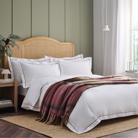 White hotel-style duvet set with grey border trim, styled on a rattan headboard bed with a burgundy plaid throw, in a sage green bedroom