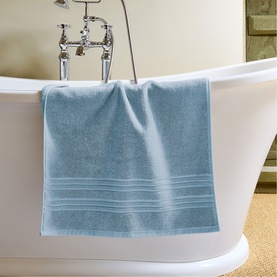 Christys Bath Towelling in Faded Denim, draped over the edge of a freestanding white bathtub in a bathroom setting.