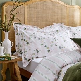Christys green floral and stripe bedding set styled on a bed with rattan headboard, paired with white vase and plant on wooden side table.
