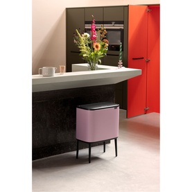 Brabantia Lilac Pink kitchen bin with pedal lid, styled in a modern dark kitchen with floral arrangement and red cabinetry
