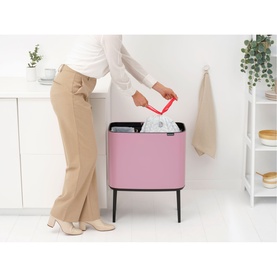 Model with Brabantia Lilac Pink bo touch bin, placing bin liner inside, styled in a modern kitchen setting with shelving.